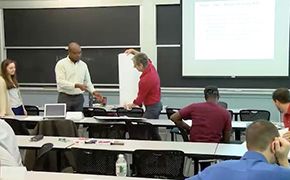 Image of instructor and students in a classroom. Two students are standing near the instructor and three are sitting at tables. A slide is projected on a pulled down screen.