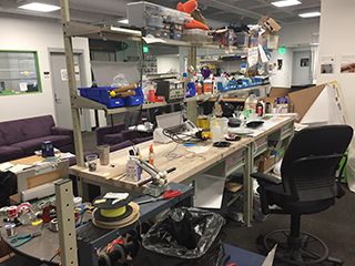 Workbench littered with pliers, wires, and bins of tools. Couches and chairs in the background.