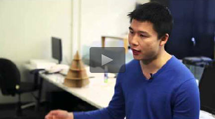 Instructor William Li sits in front of a long white table. He’s gesturing with one arm. There are two computer monitors on the table, along with a three dimensional object constructed out of cardboard.