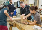 A greying-haired google-wearing gentleman helps a couple of students screw an object into a wooden block.