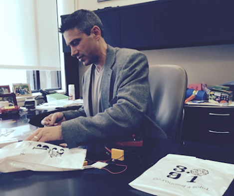 A man sitting at a desk, on which plastic bags and assorted small objects are lying.