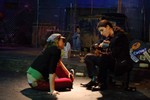 A man with a pony-tail plays an acoustic guitar for a girl, while a man sits next to a dumpster in the background.