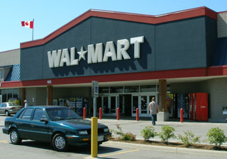 A small blue car is parked in front of a Wal-Mart entrance.