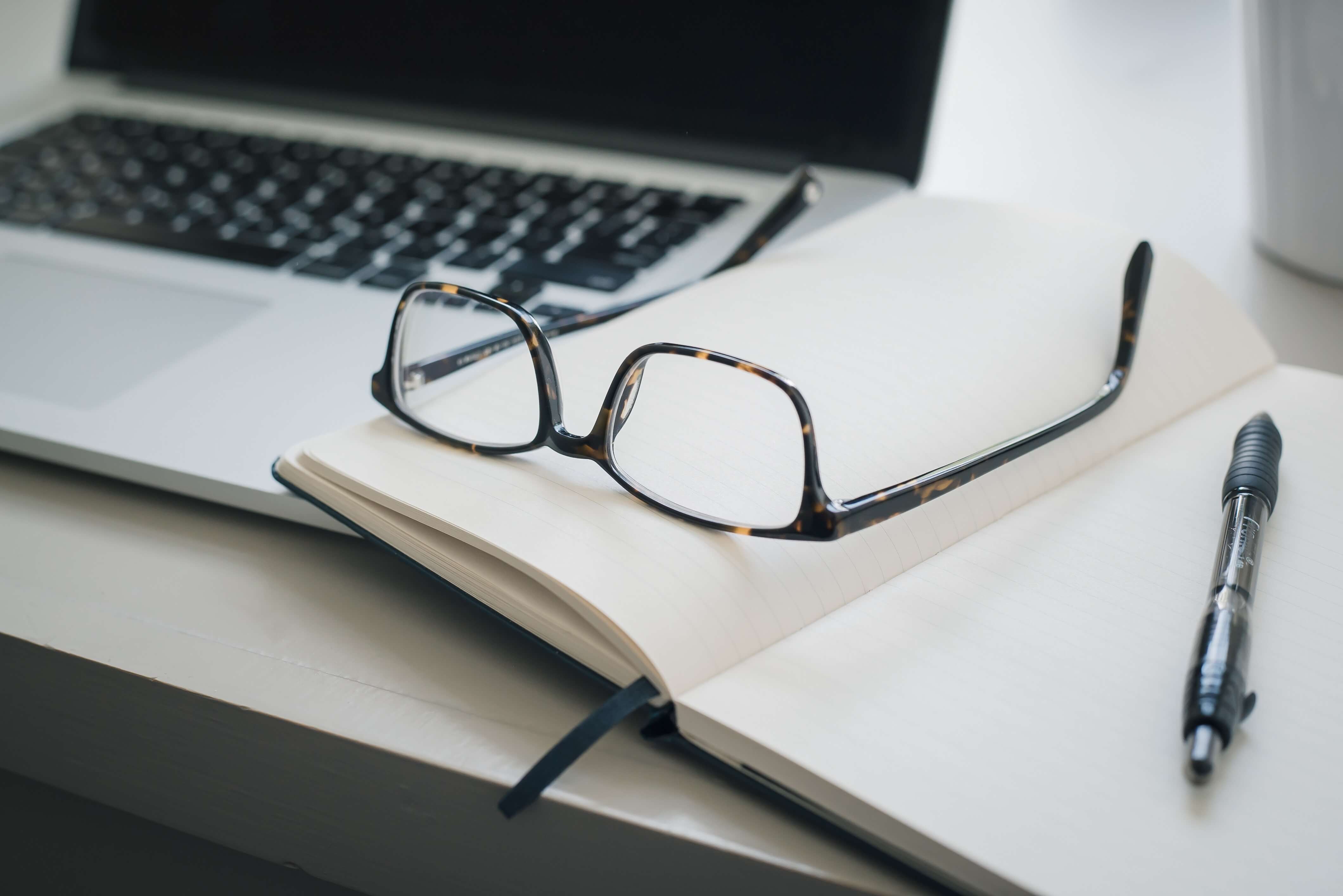 A photo of an open laptop and notebook with a pen and a pair of glasses resting on them.