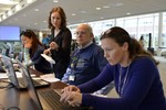 Three volunteers sit in front of laptops, while being instructed by a young woman.